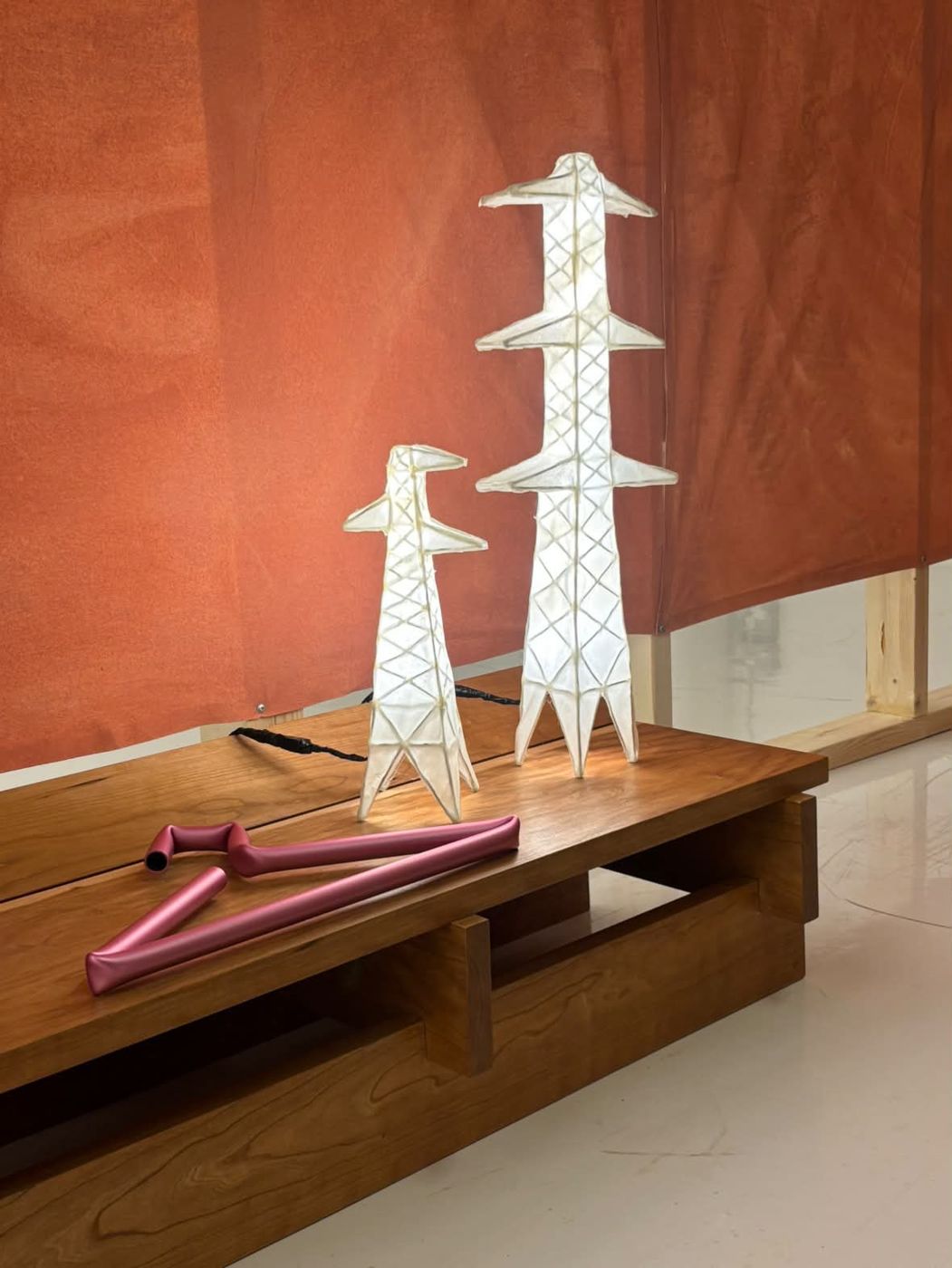 Two Pylon Lamps glowing on a cherry wood bench in front of a rust-orange fabric wall