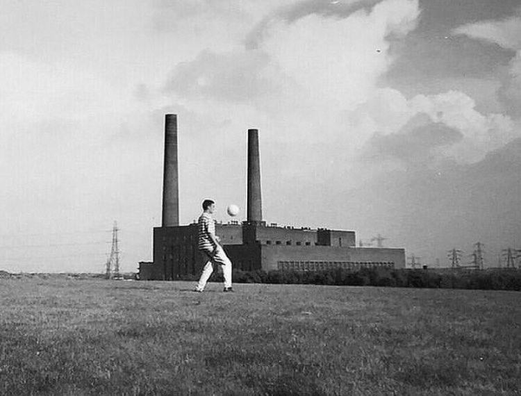 Black and white photograph of a man kicking a football in a field in front of a two-chimney power station