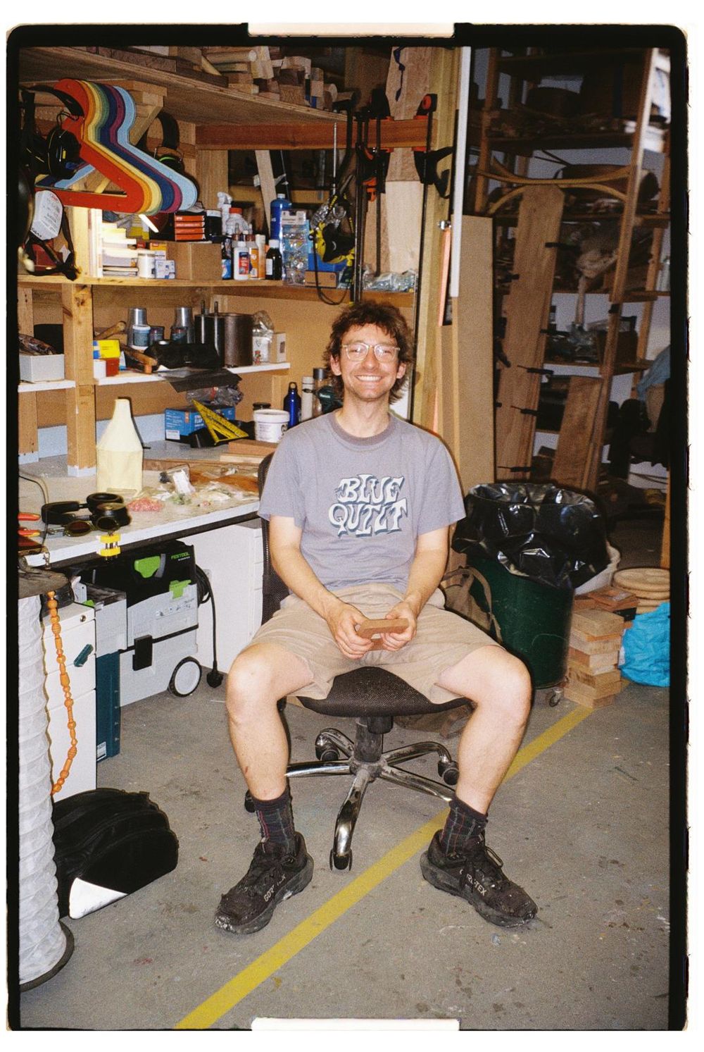 Sam Braid smiling, sat on a stool in his workshop surrounded by timber racks, clamps and tools